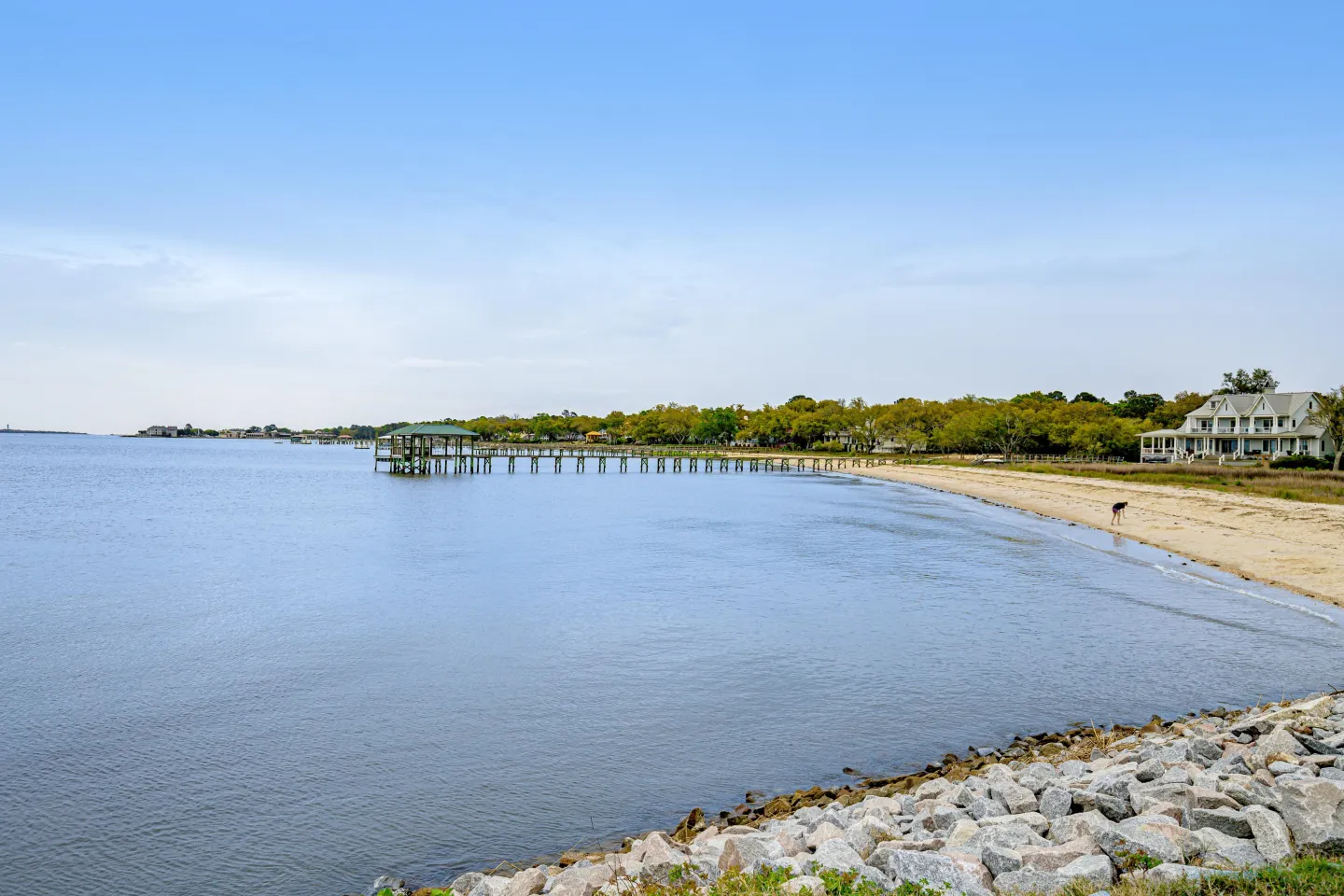 Wide view of a calm bay shoreline with a sandy beach, a long wooden dock with a covered pier head, and a house in the distance.