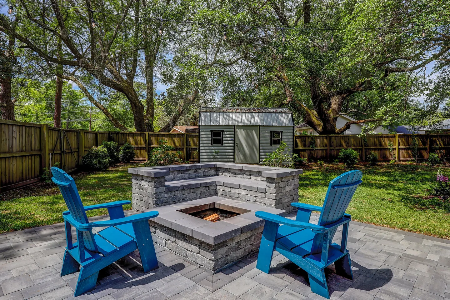 Fenced backyard featuring a stone fire pit, built-in bench seating, two bright blue Adirondack chairs, and a storage shed in the background.