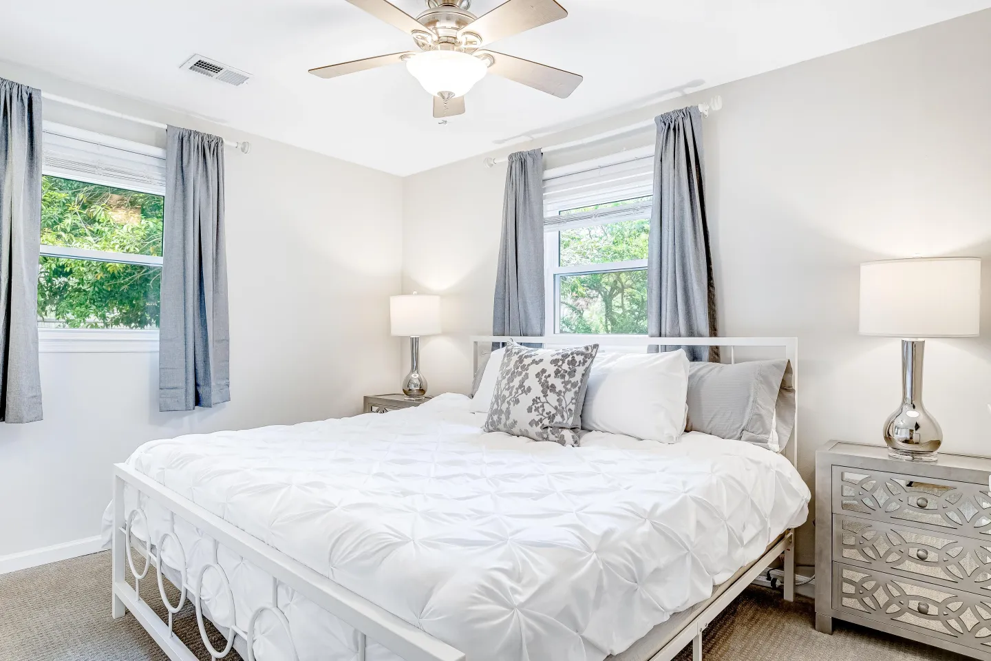 Bedroom with a white metal bed, plush white bedding, mirrored nightstands with lamps, grey curtains, and a ceiling fan.