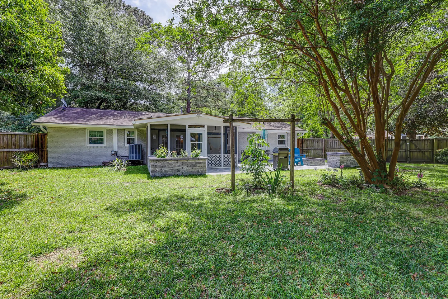 Rear exterior of a single-story brick home featuring a fenced green backyard, screened-in porch, open patio, and mature trees.