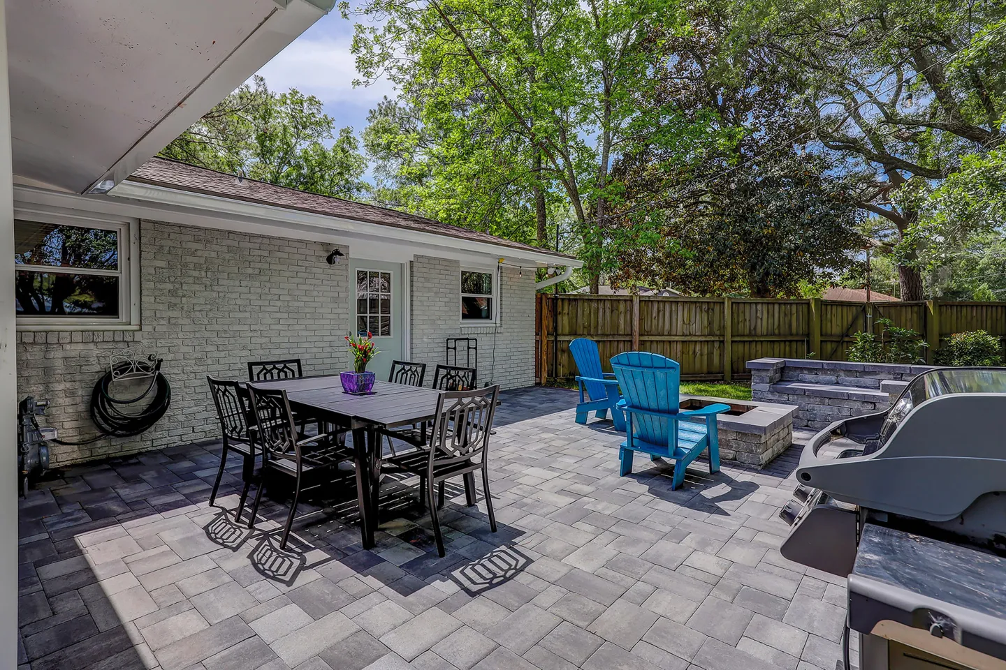Outdoor paved backyard patio with a dining table and chairs, blue Adirondack chairs by a fire pit, and a barbecue grill next to a brick house.