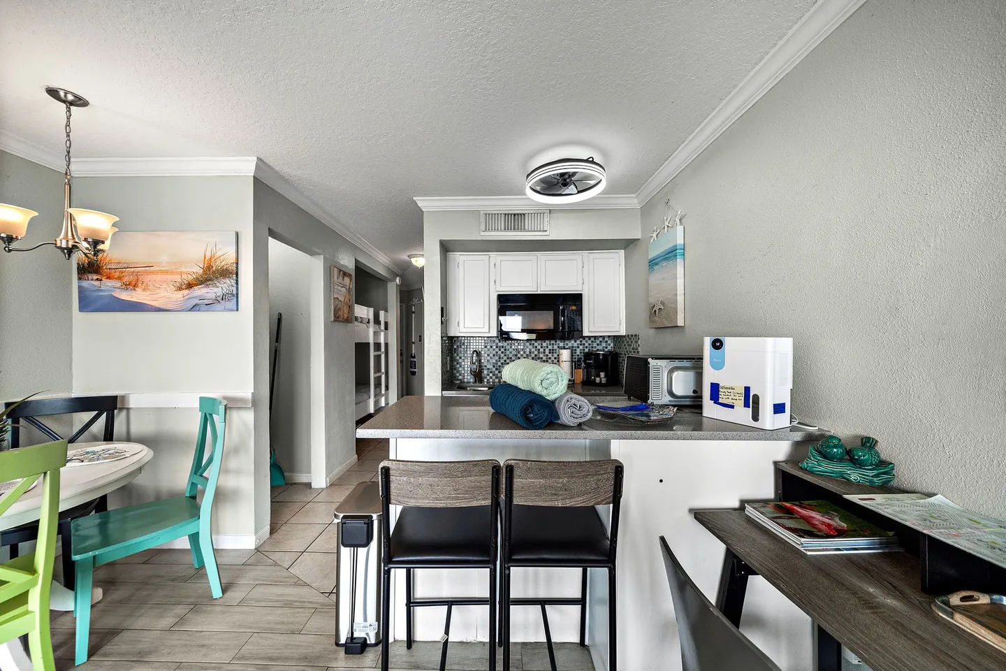 Open dining area and kitchen with breakfast bar stools, light-colored tile floors, and a hallway leading to bunk beds.