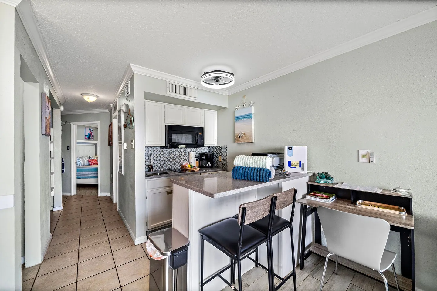 Compact kitchen and dining counter with barstools, next to a small desk, with a hallway leading to a bedroom.