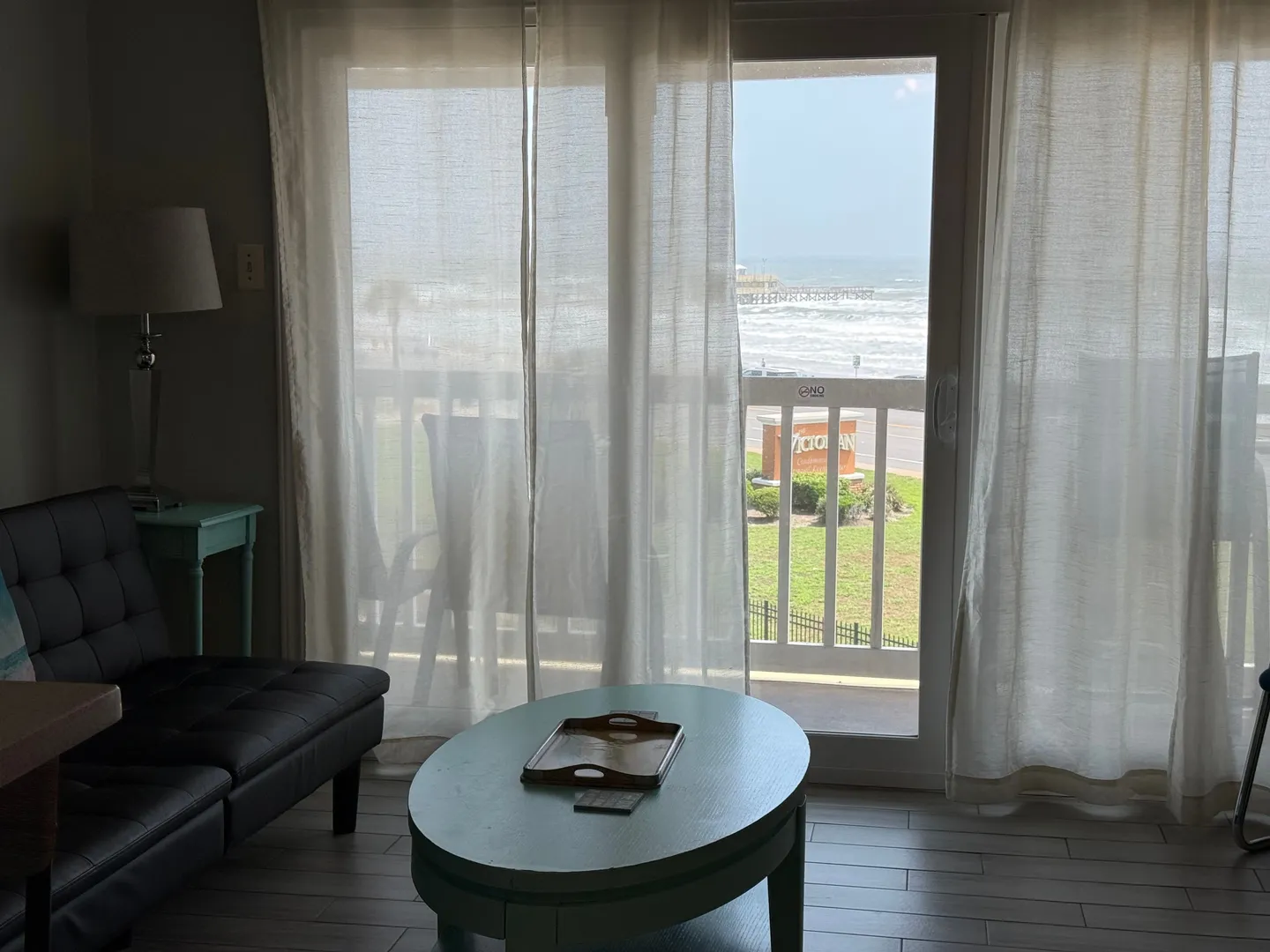 Living area with an oval coffee table, dark sofa, and sheer curtains framing a sliding glass door with a view of a balcony and the ocean.