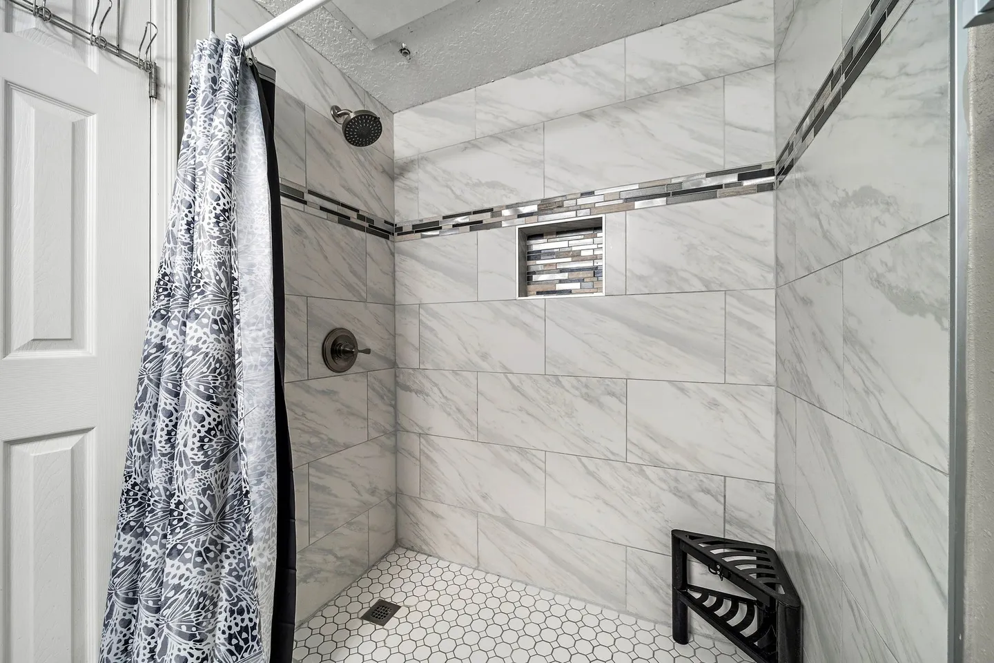 Walk-in shower with marble-look wall tiles, mosaic accent border, shower niche, hexagonal floor tile, and a corner stool.