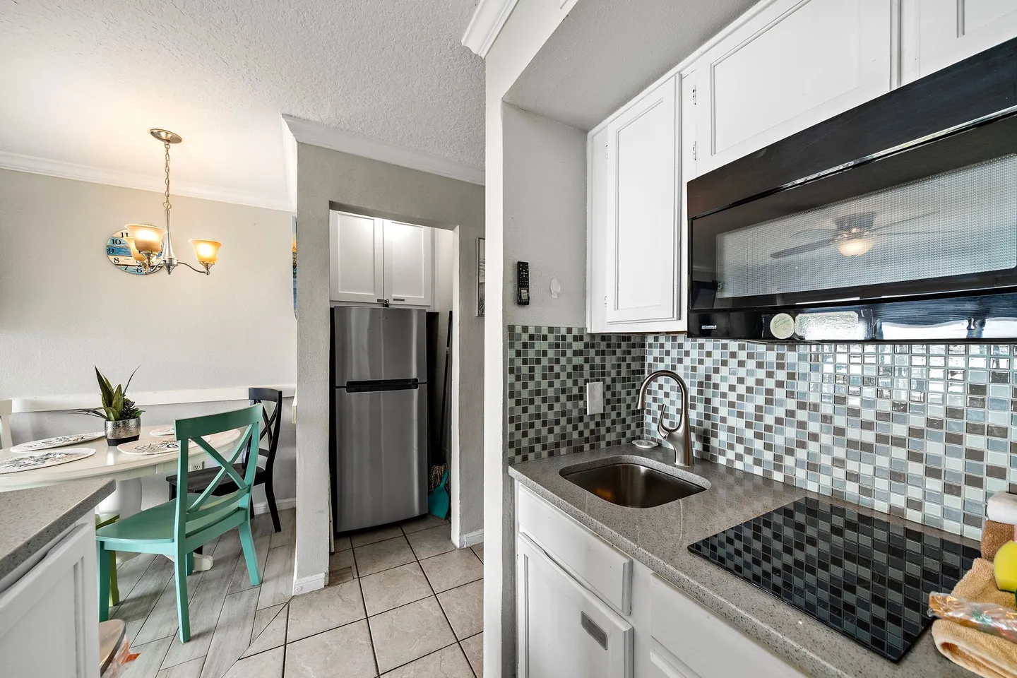Kitchenette with white cabinetry, glass tile backsplash, countertop, sink, cooktop, and view into an adjacent dining area.