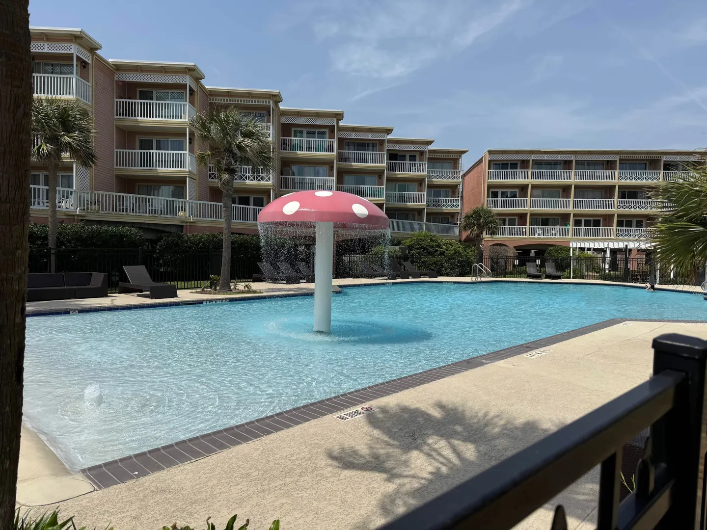 Outdoor swimming pool with a mushroom-shaped water fountain feature in front of multi-story condominium buildings.