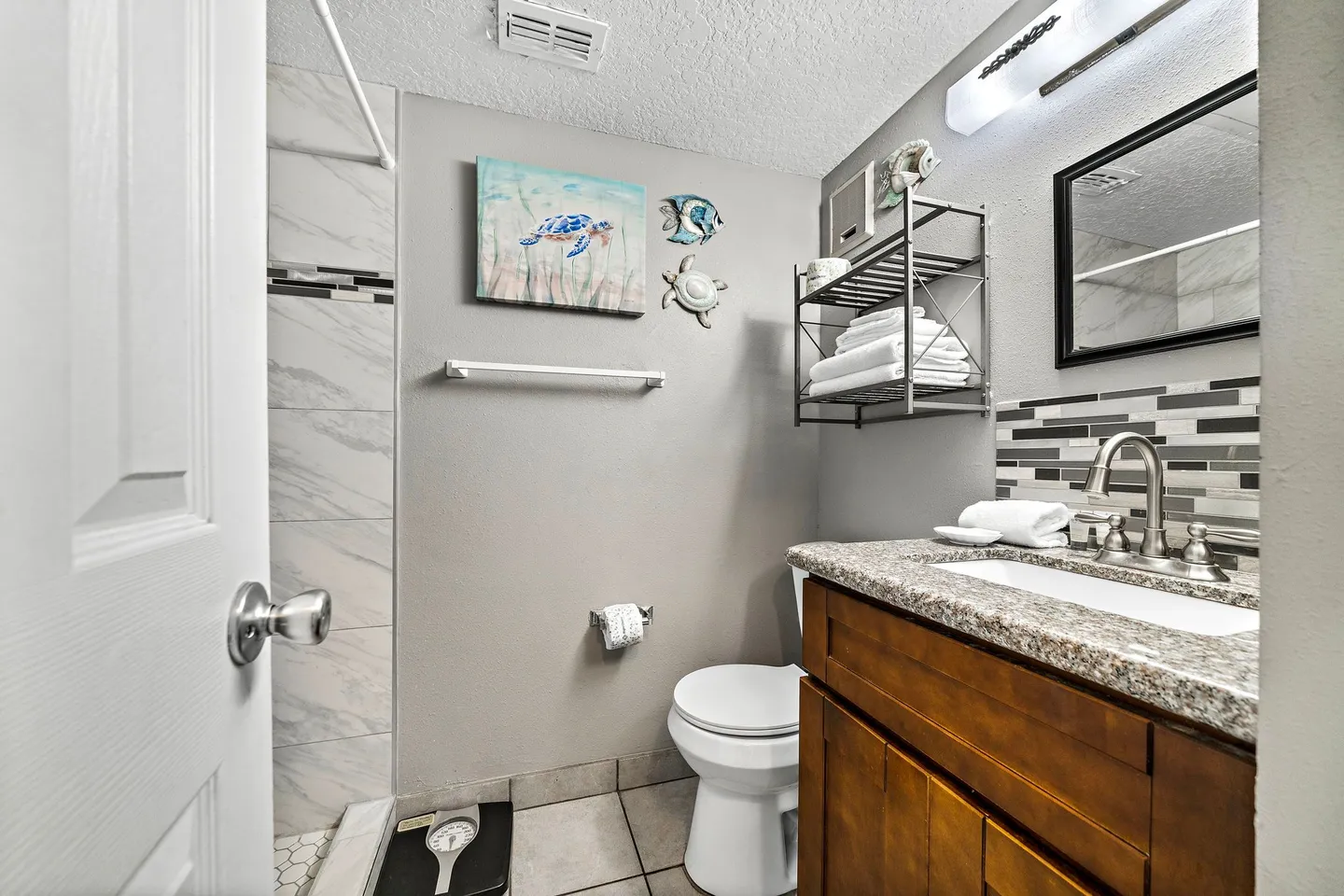 Bathroom featuring a dark wood vanity with a speckled countertop, a toilet, sea-life wall decor, and a tiled walk-in shower on the left.