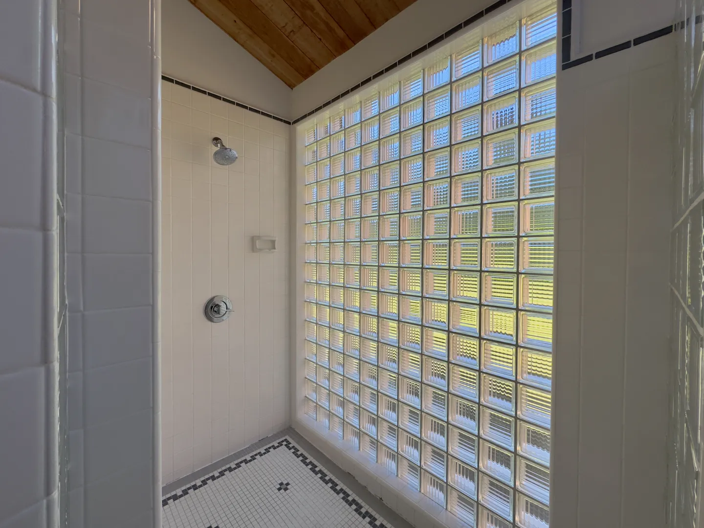 Walk-in shower featuring a large glass block wall, white tiled walls, patterned tile floor, and a sloped wood ceiling.