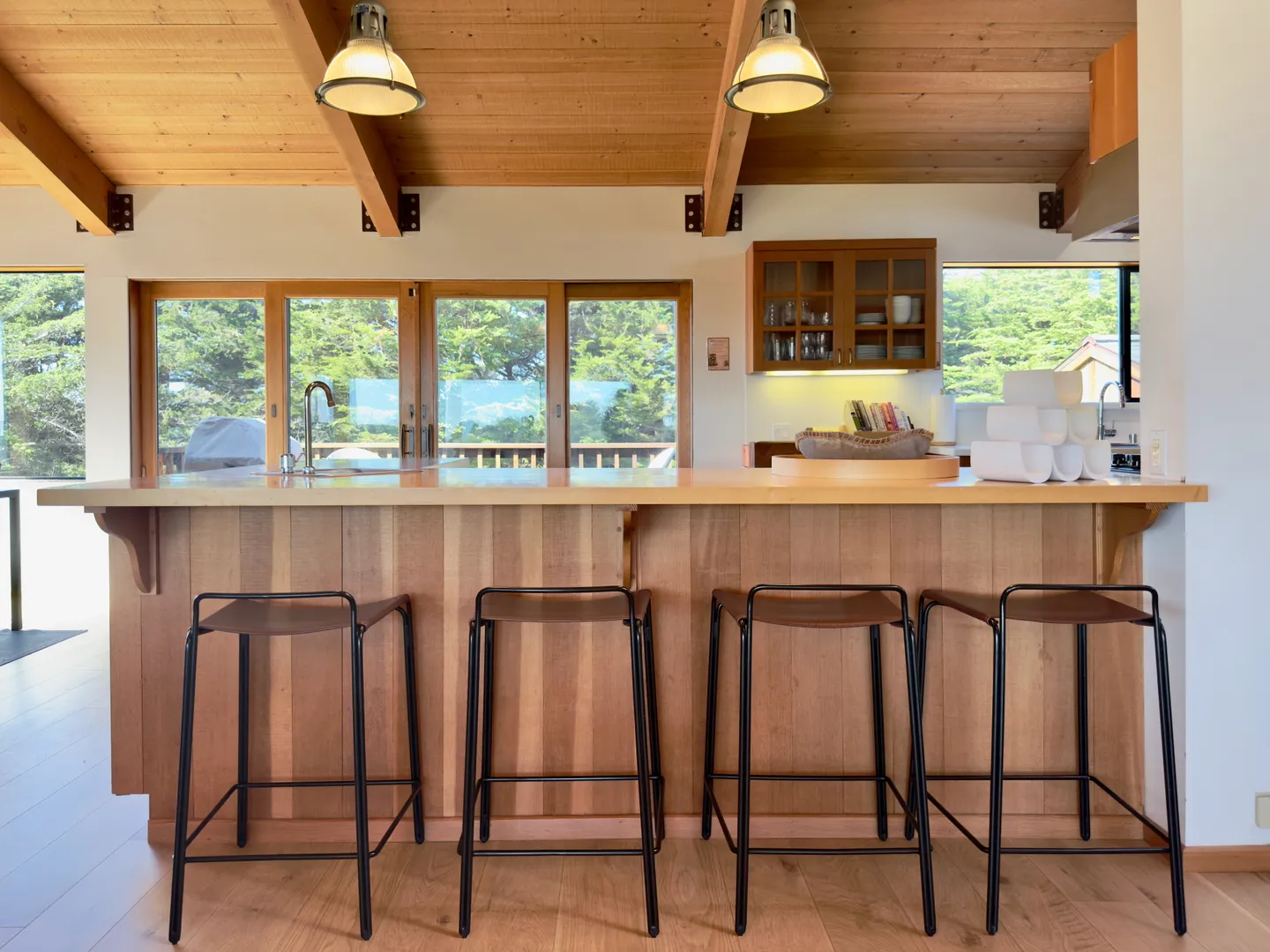 A kitchen island with four barstools and a wood-paneled base under a wood-beamed ceiling with sliding glass doors in the background.