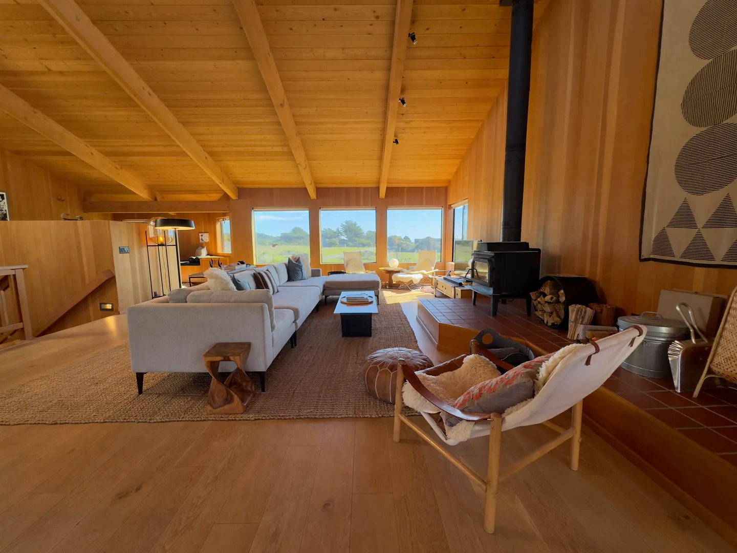 Living room with wood paneled walls, vaulted beamed ceiling, large sectional sofa, wood burning stove, and picture windows.