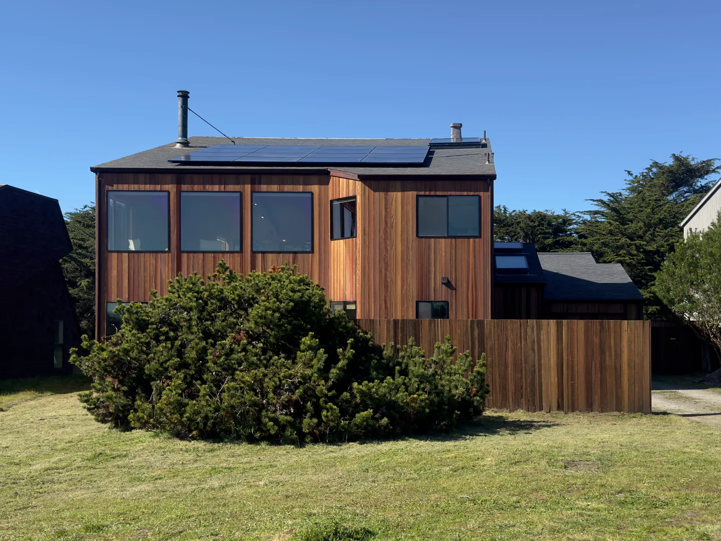 Exterior of a two-story home with vertical wood siding, solar panels on the roof, a wooden privacy fence, and a grassy yard under a clear blue sky.
