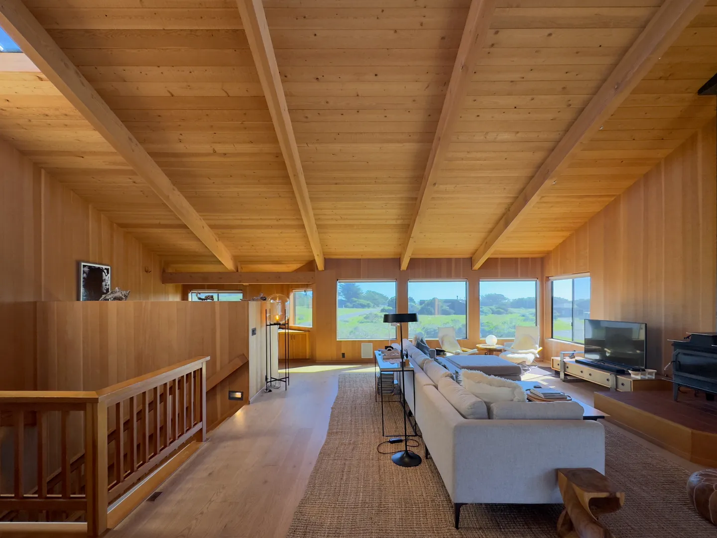 Open living area with wood-paneled walls and ceiling, large windows looking out to coastal scenery, and a central seating arrangement with a sofa and television.