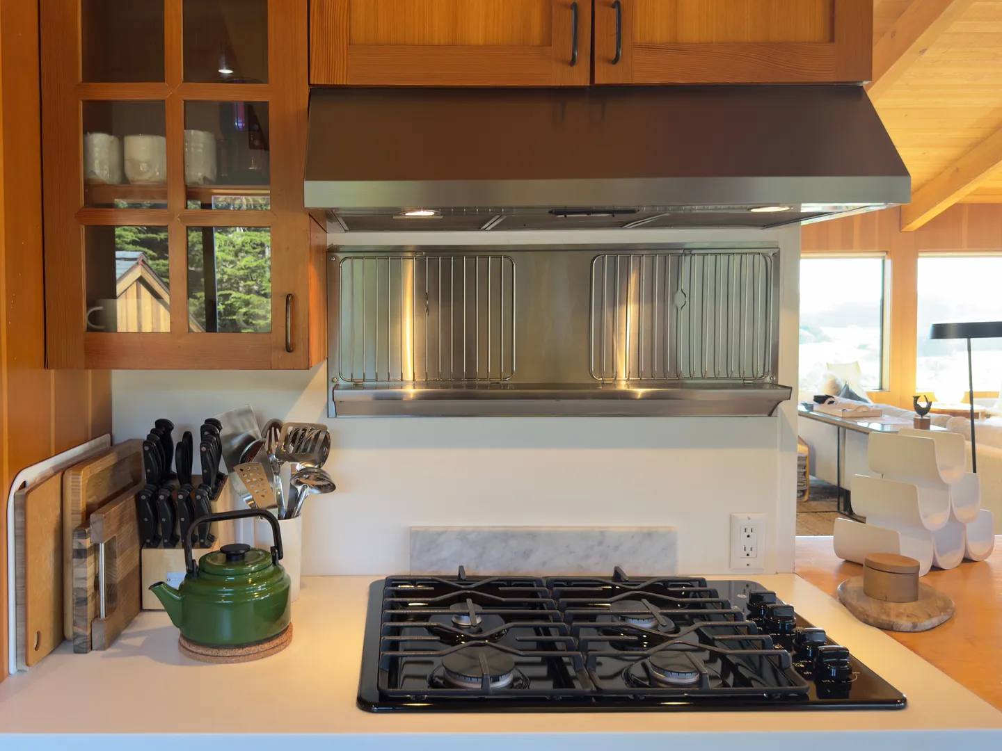 Kitchen counter area with a built-in black gas cooktop beneath a stainless steel vent hood, alongside wooden cabinets and cooking accessories.