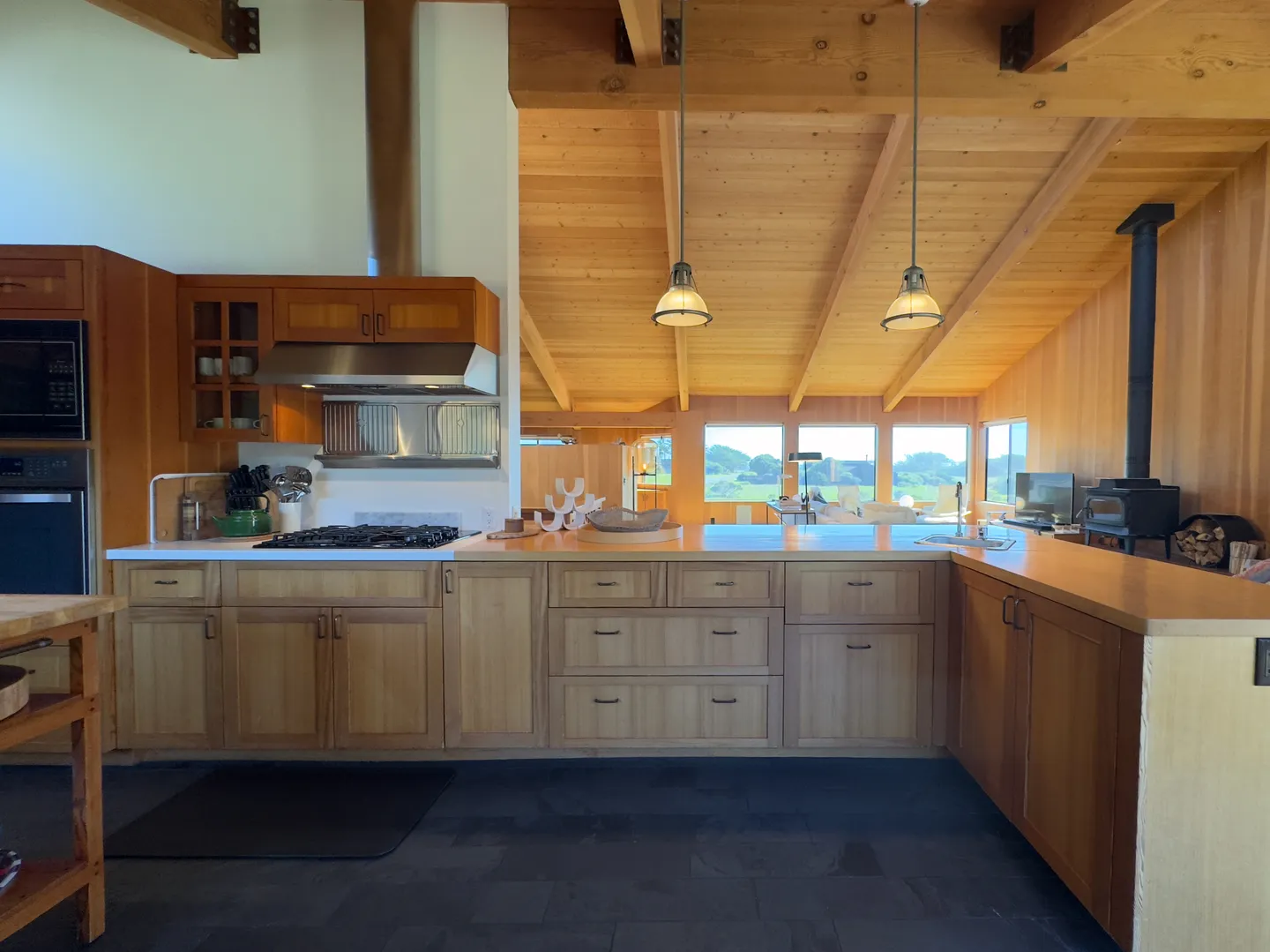 Open-concept kitchen with light wood cabinetry, exposed beam wood-paneled ceiling, and views into an adjacent living area with windows.