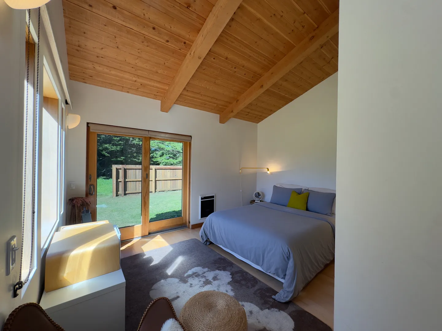 Bedroom with vaulted wood plank ceiling, made bed, area rug, and sliding glass door leading to a fenced yard.