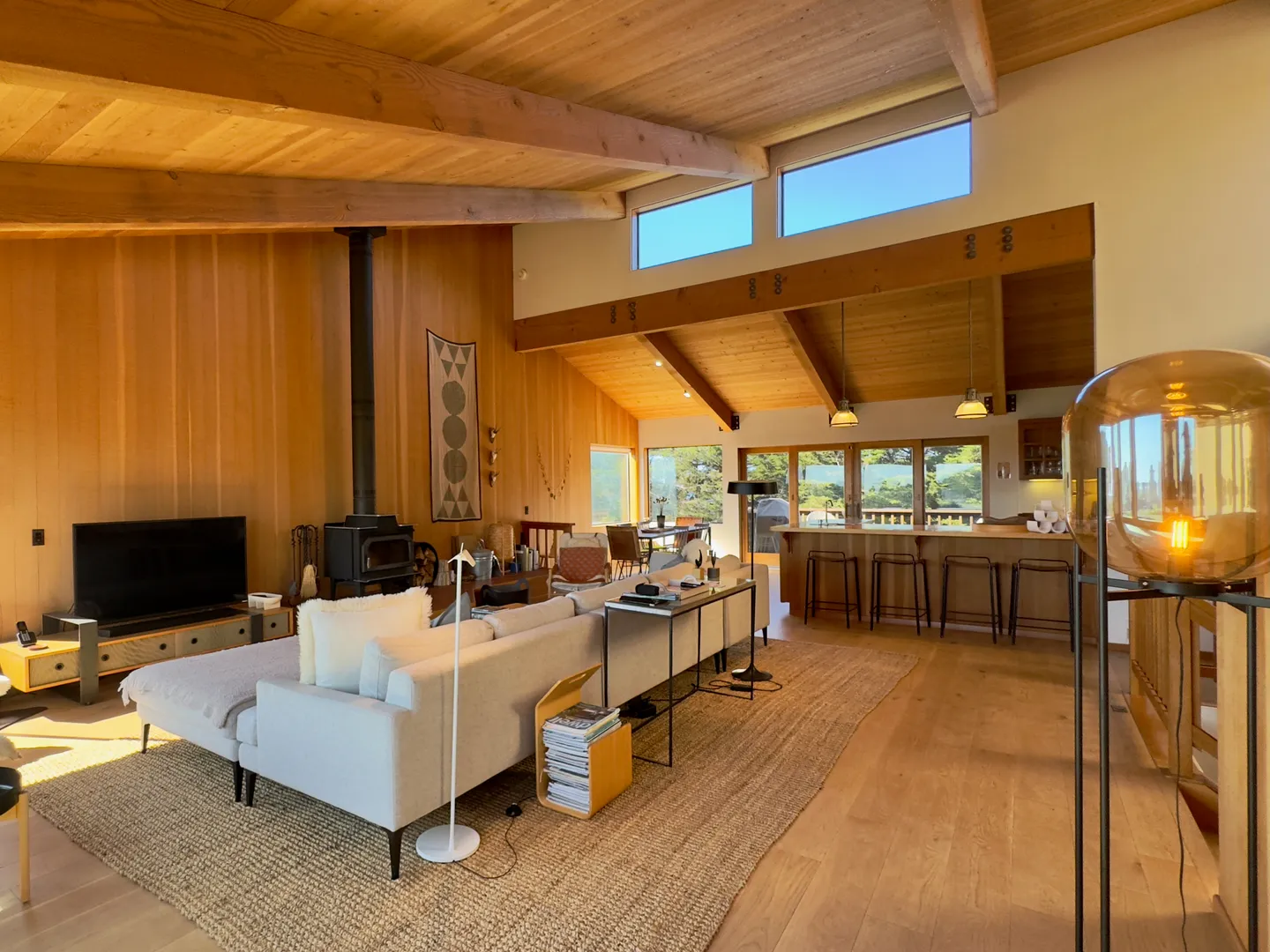 Open-concept living room with vaulted wood-beamed ceilings, wood-paneled walls, a sectional sofa, a wood-burning stove, and a view into the kitchen with breakfast bar seating.