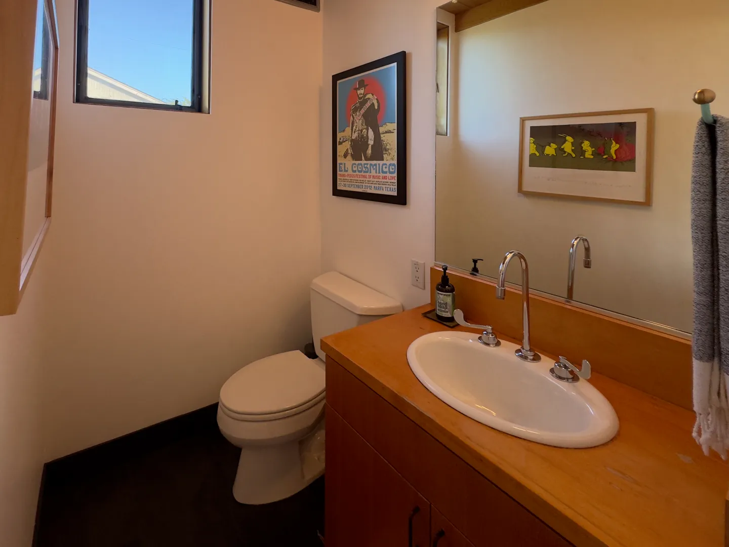Bathroom with a wood vanity, white oval drop-in sink, toilet, framed art, and a small high window.