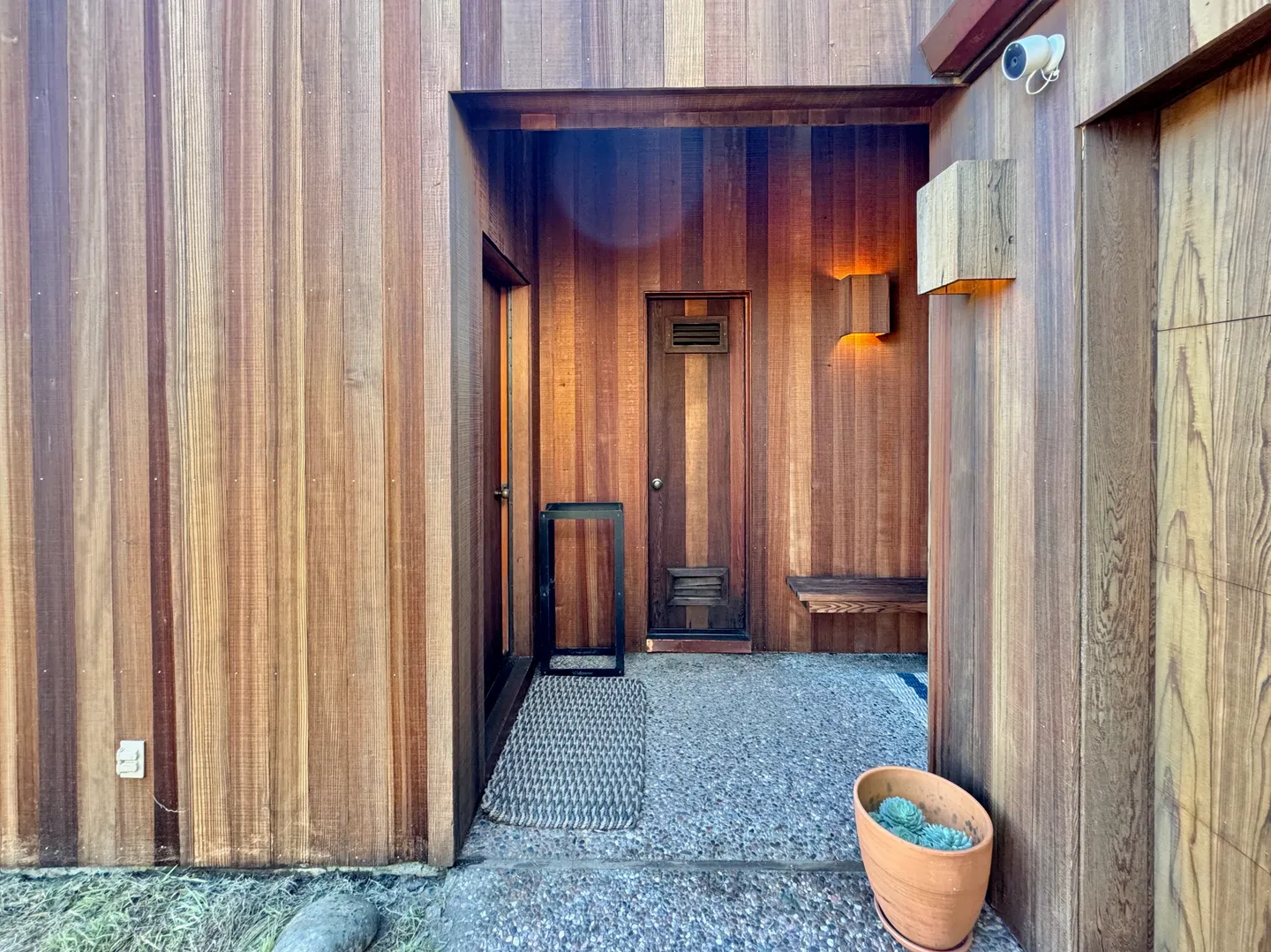 Covered outdoor entryway with vertical wood paneling, a gravel floor, warm lighting, and a front door.