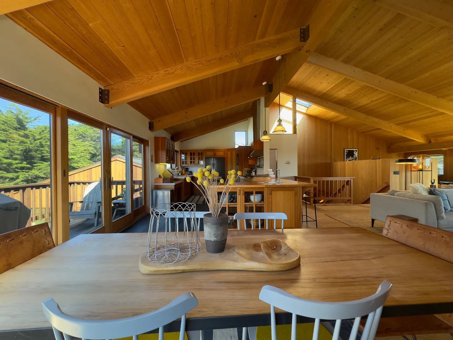 Open-concept dining, kitchen, and living area beneath vaulted wood-paneled ceilings, viewed from behind a dining table.