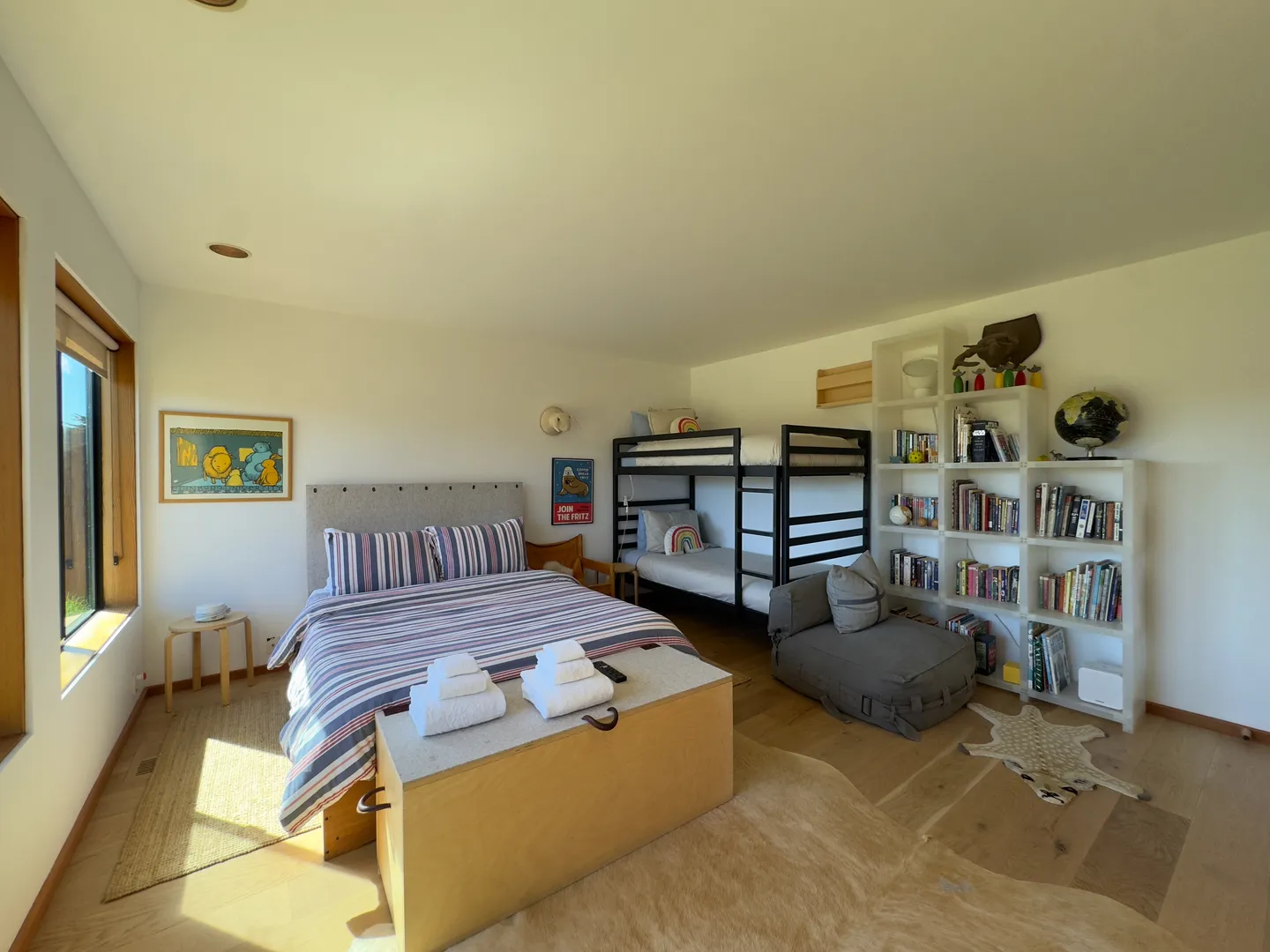 Bedroom with a bed made with striped bedding, a metal bunk bed, a tall white bookcase filled with books, and a low storage chest.