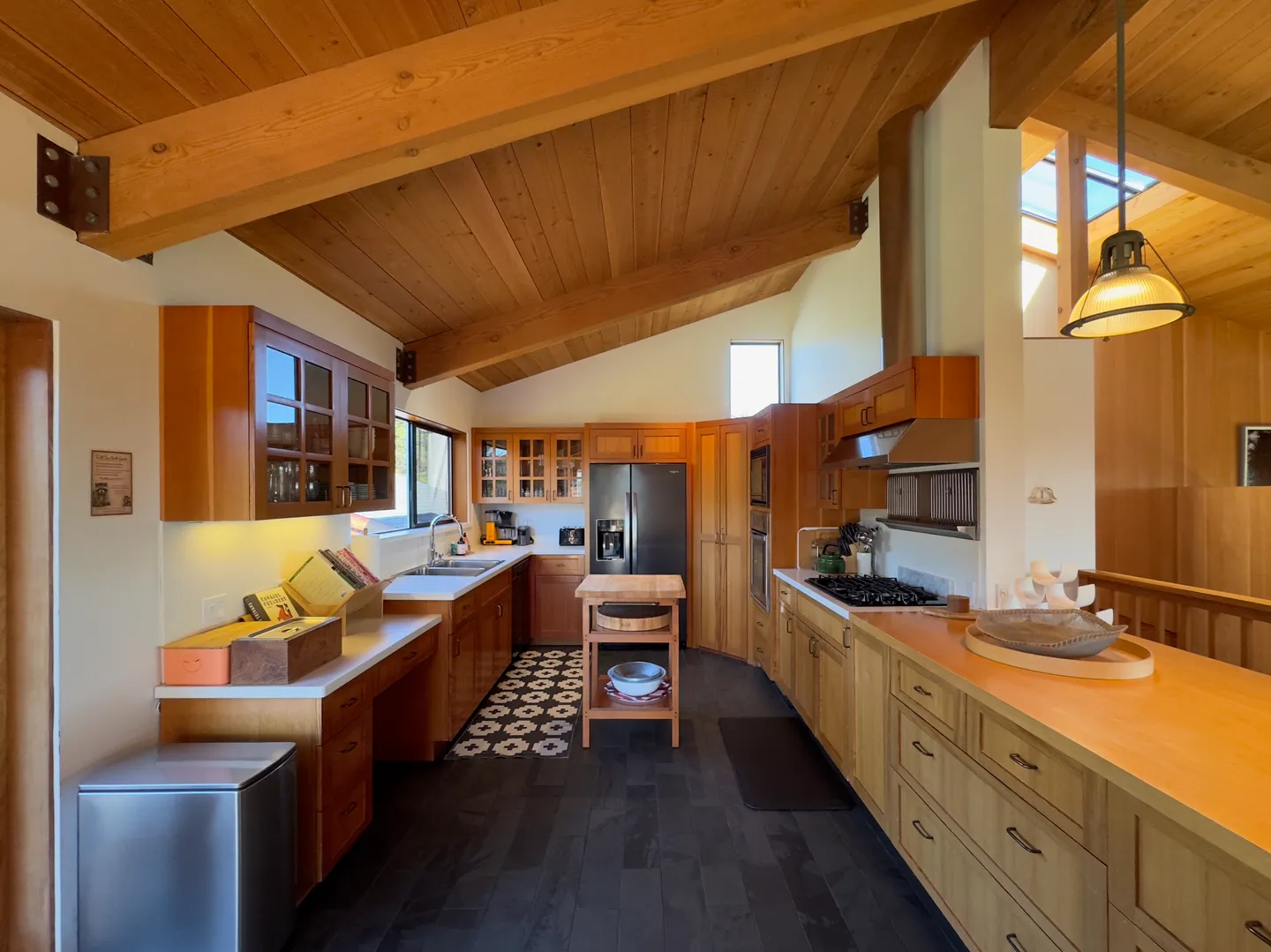 Spacious kitchen featuring angled wood-paneled ceilings, exposed wood beams, light wood cabinetry, and dark tiled flooring.