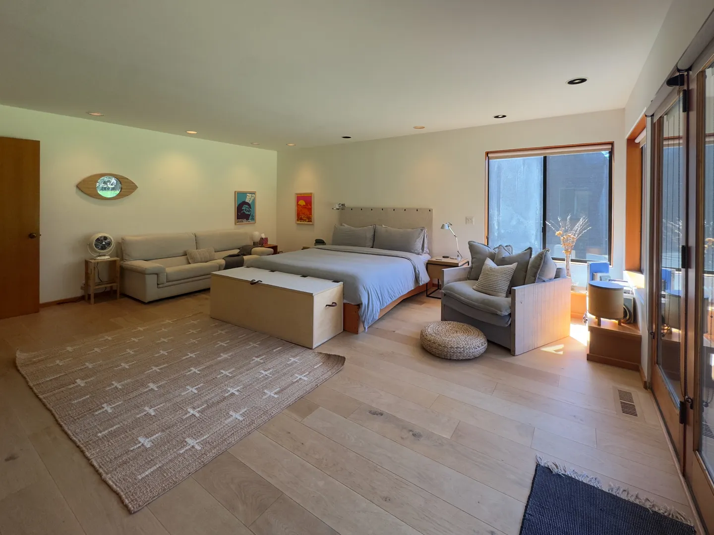 Spacious bedroom with a made bed, couch, armchair, wooden bench, and light wood flooring.