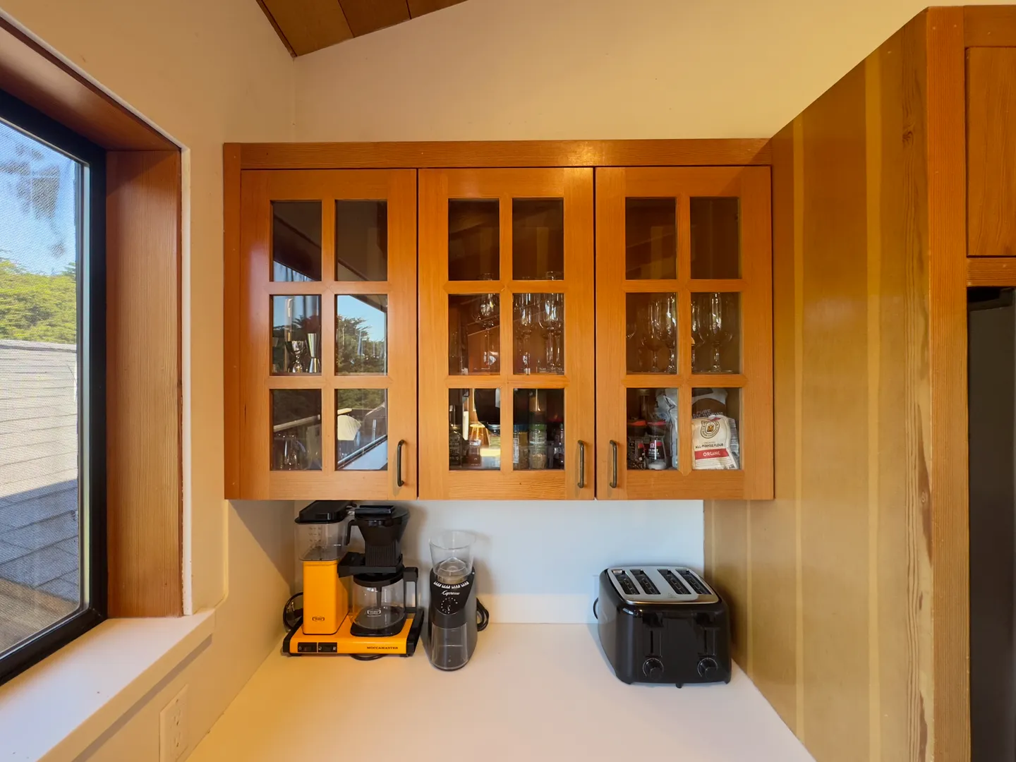 Kitchen counter nook with wooden glass-front upper cabinets, a window to the left, a drip coffee maker, coffee grinder, and a four-slice toaster.