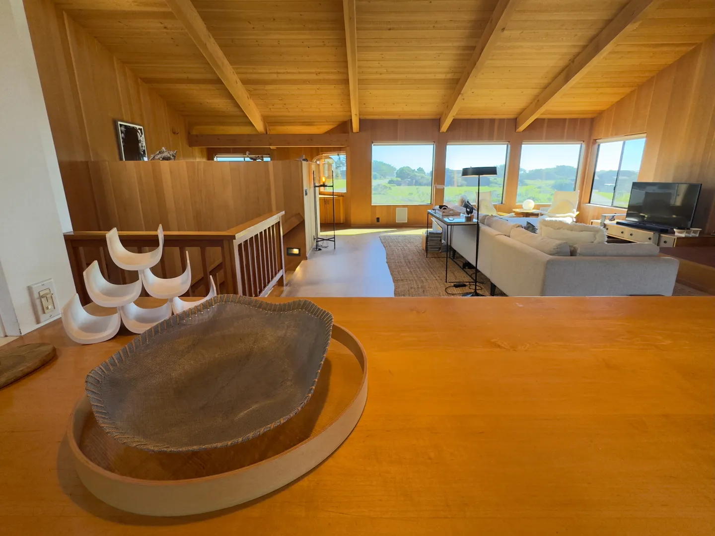 Elevated view over a wooden counter looking down into a wood-paneled living room with a sectional sofa and large picture windows.