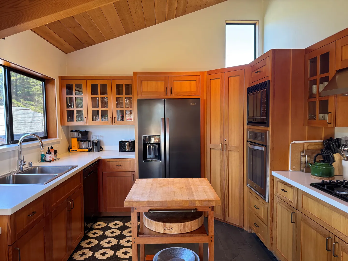 Kitchen with wood cabinetry, white countertops, built-in ovens, a stainless steel refrigerator, and a butcher block island.