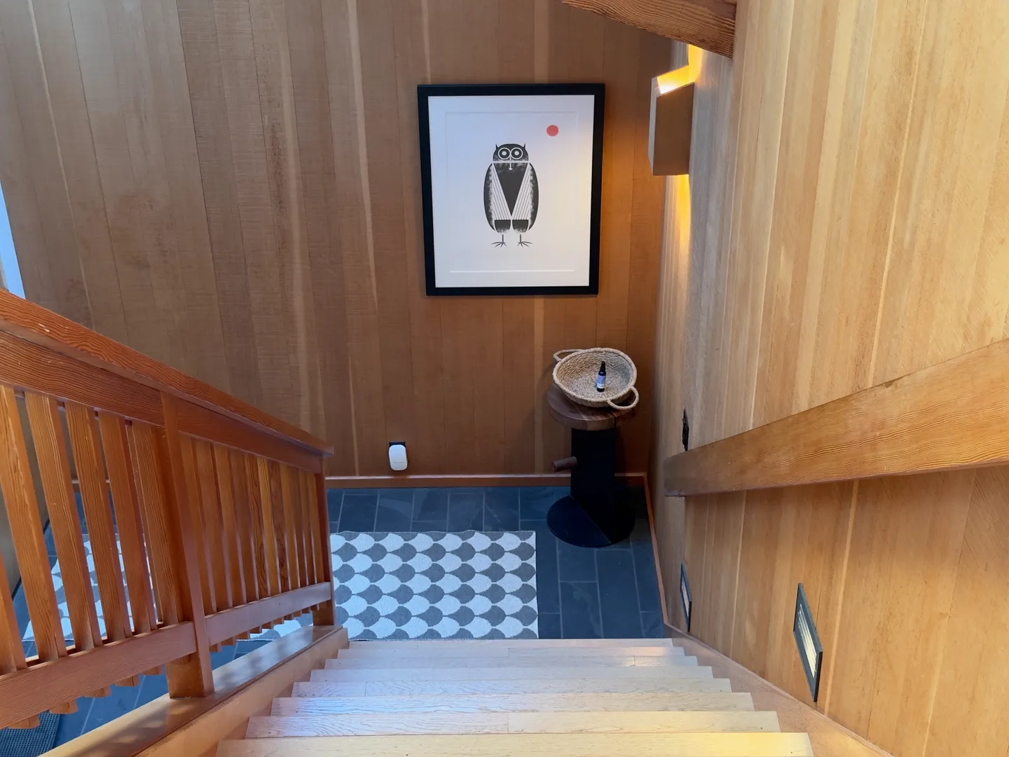 Looking down a light wood staircase toward a tiled landing with wood-paneled walls, an owl art print, and a patterned rug.
