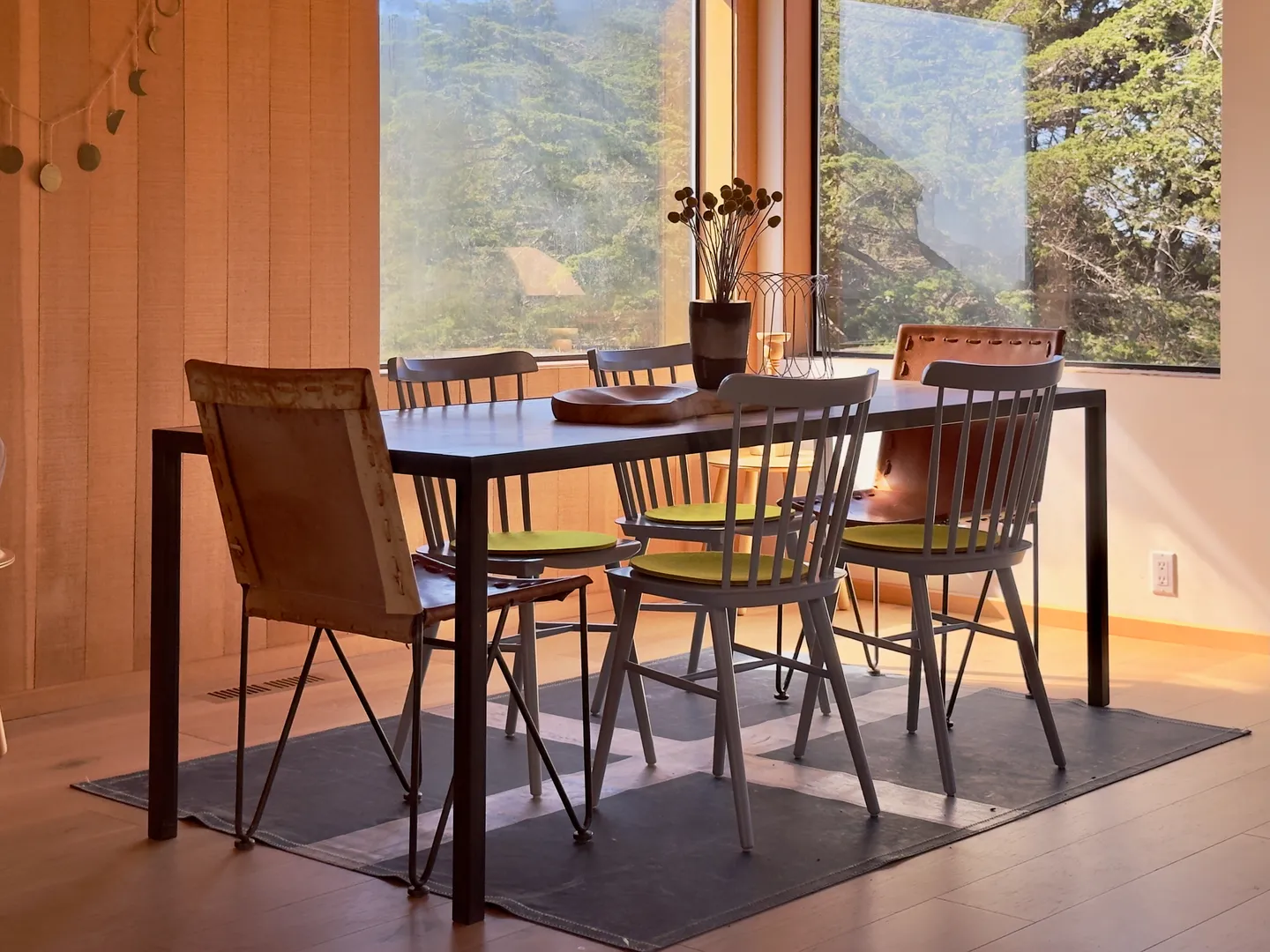 Dining table and chairs set next to large windows overlooking green trees.