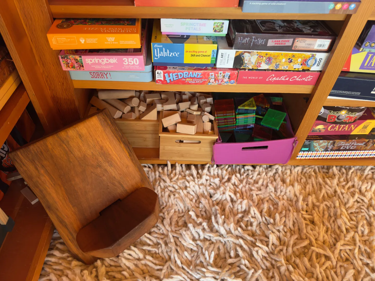 Low wooden shelf stocked with board games, puzzles, wooden blocks, and magnetic tiles on a textured carpet.