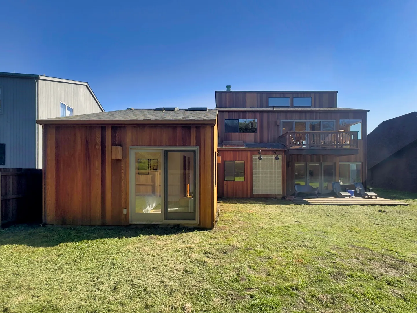 Rear exterior view of a multi-story home with natural wood siding, an adjacent studio structure, and a grassy backyard under a clear blue sky.