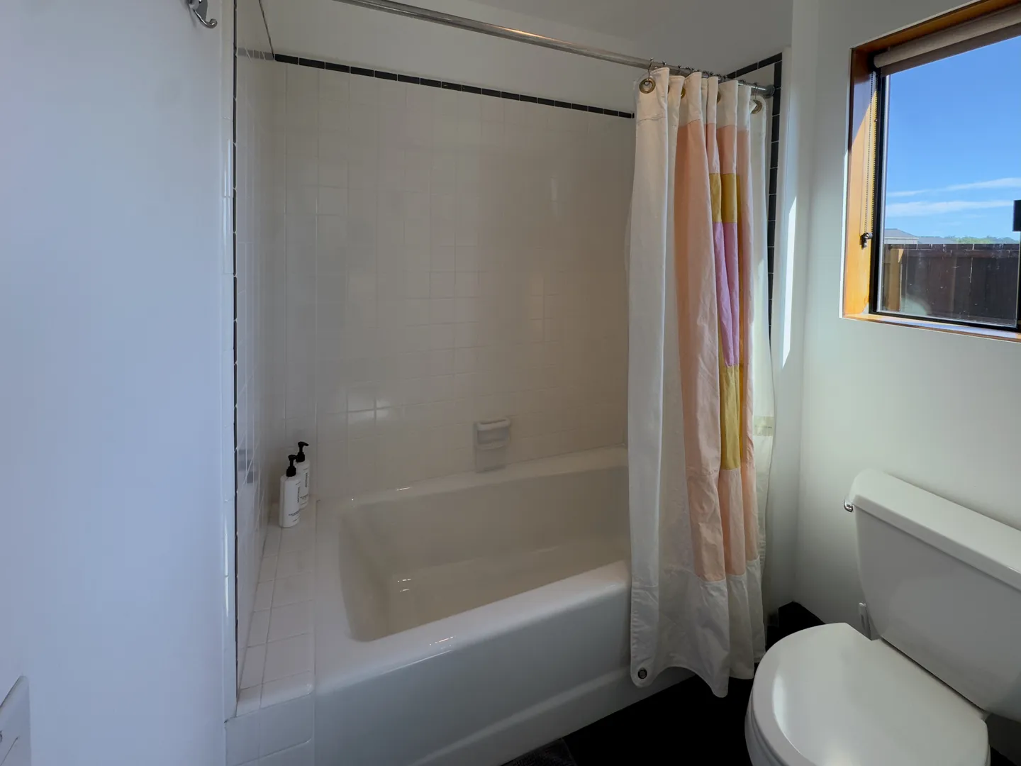 Bathroom with a white bathtub and tiled shower surround next to a toilet and a window.