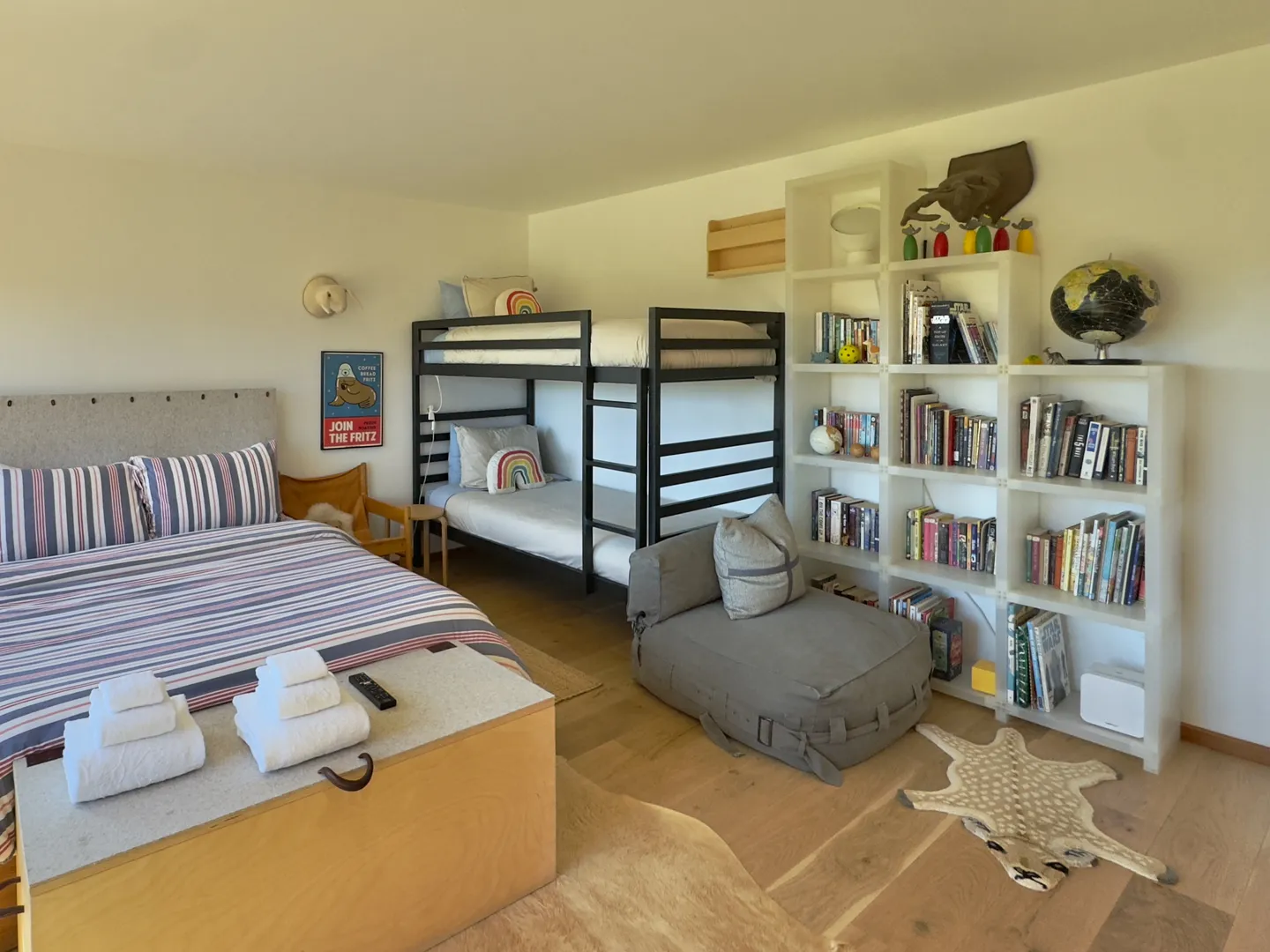 Bedroom featuring a double bed, dark metal bunk beds, a large white bookshelf filled with books, and a low lounge chair.