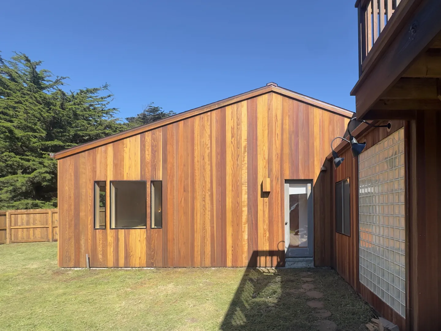 Exterior view of a home with vertical wood panel siding, a grassy yard, and a glass-block wall feature under a blue sky.