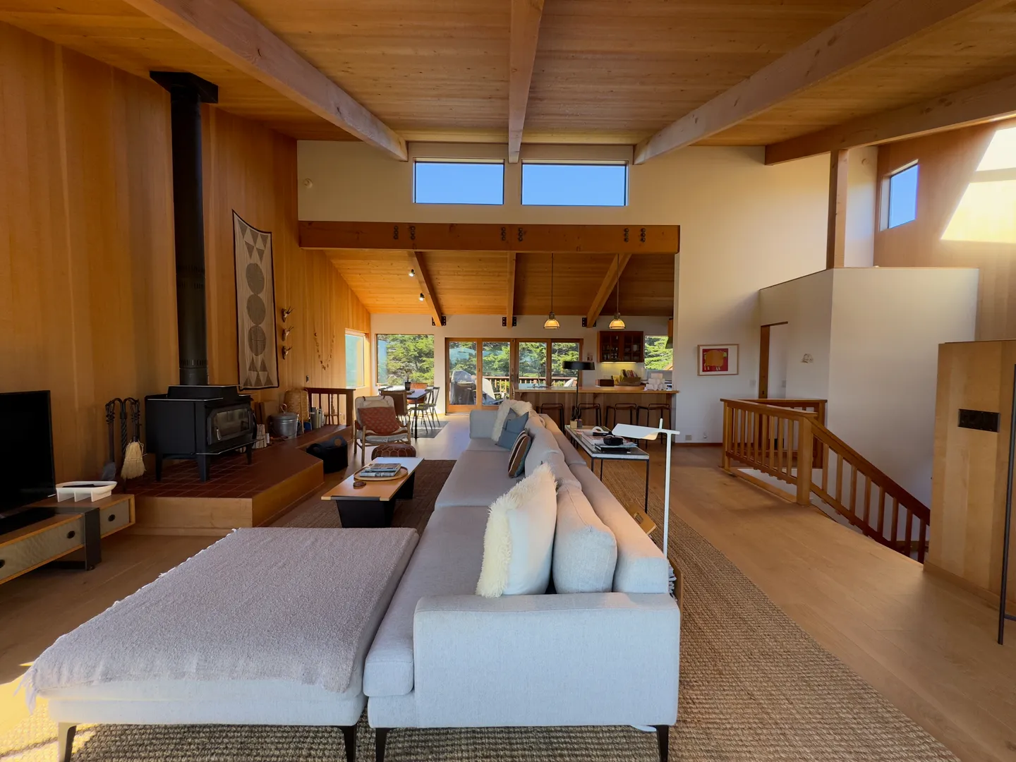 Open-concept living room with wood-paneled walls, exposed ceiling beams, a long sectional sofa, a wood-burning stove, and a view into the kitchen.
