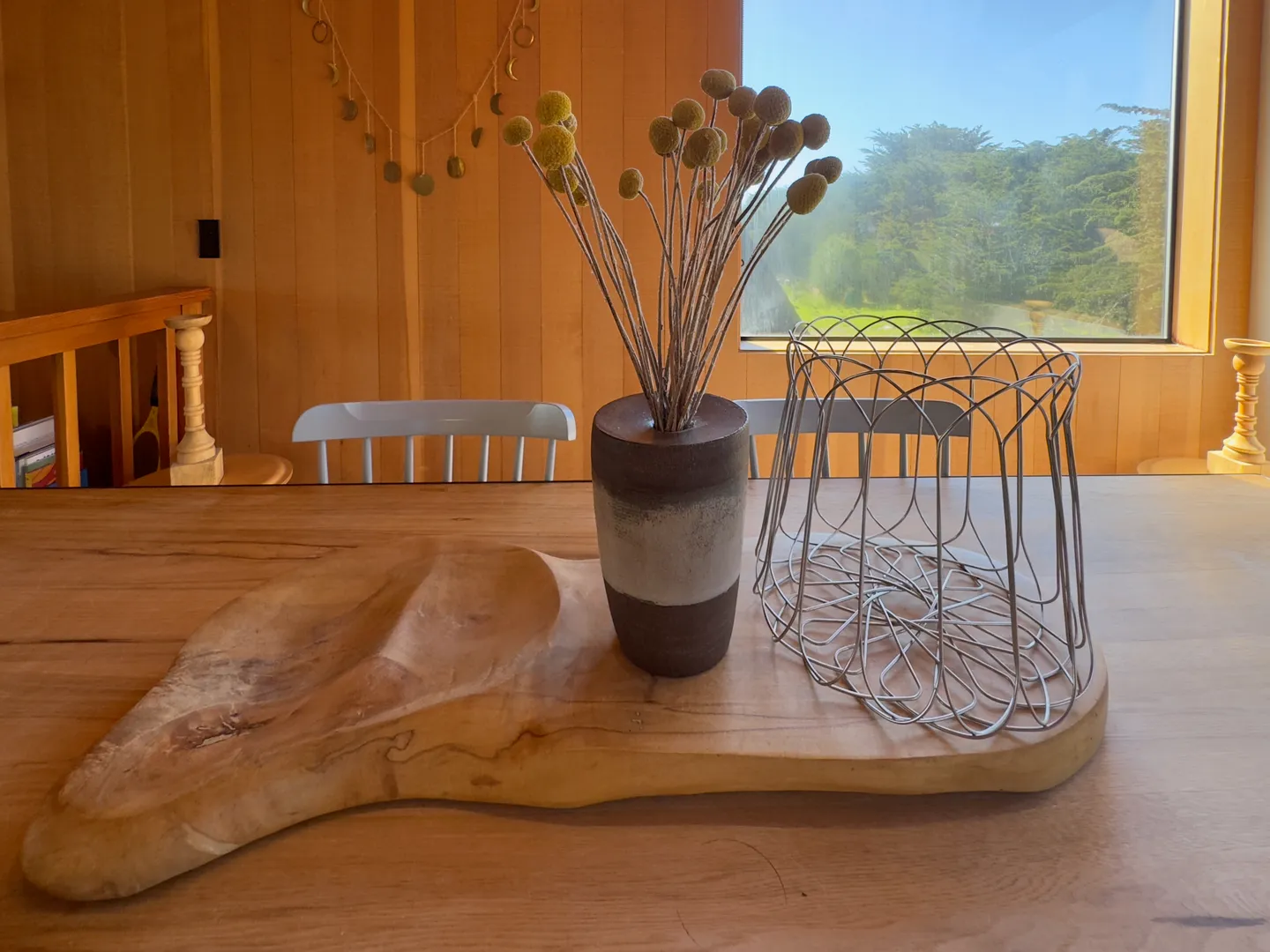 A close-up view of a dining table centerpiece featuring a carved wood board, vase with dried round seed heads, and a wire basket against wood-paneled walls.