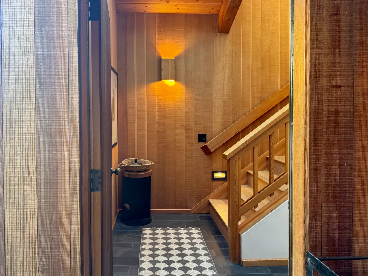 Entryway viewed through an open doorway, featuring wood-paneled walls, slate tile flooring, a geometric rug, and a wooden staircase.