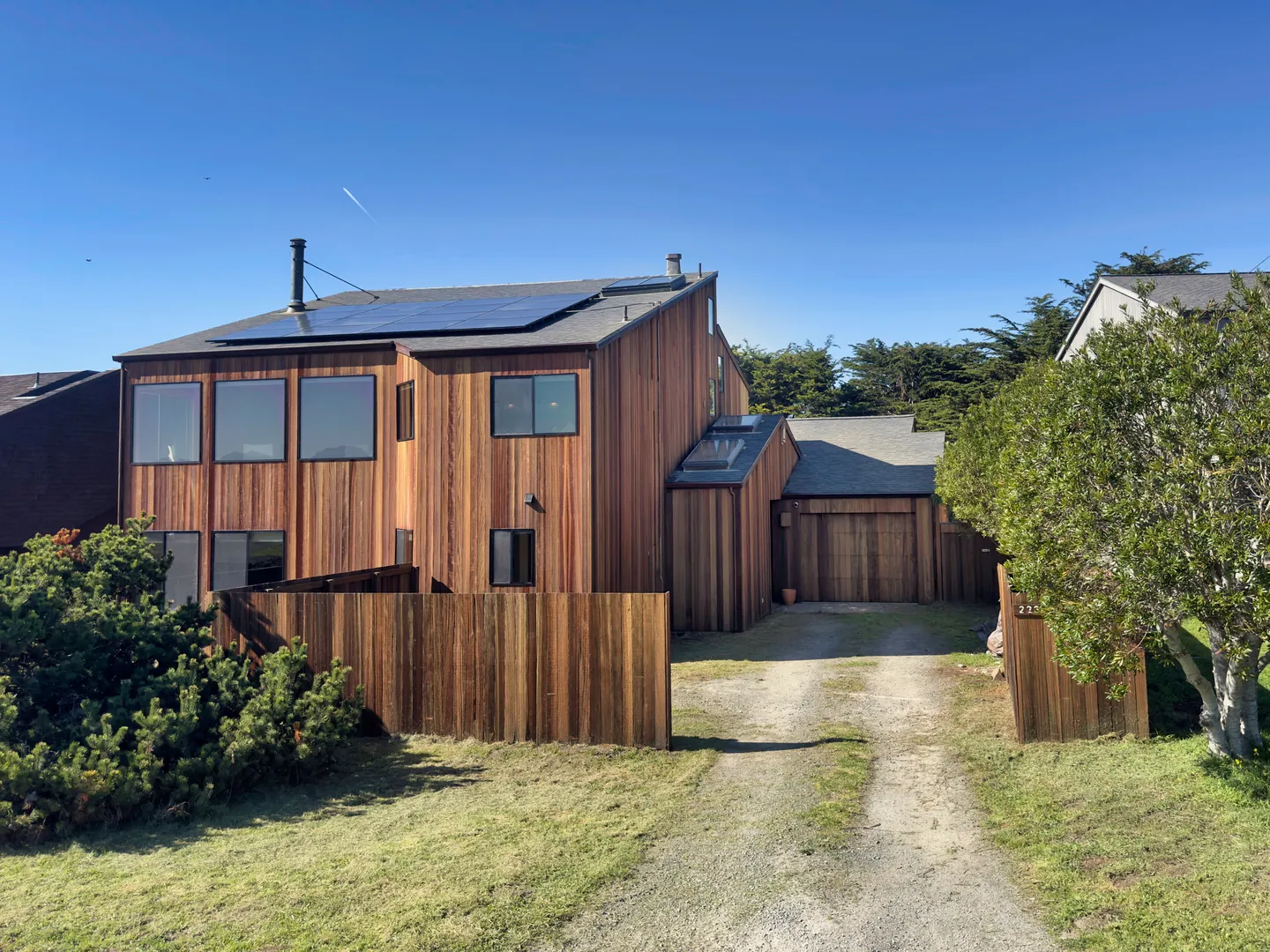 Exterior view of a multi-story wood-sided house with a gravel driveway, wood fence, and solar panels on the roof under a clear blue sky.
