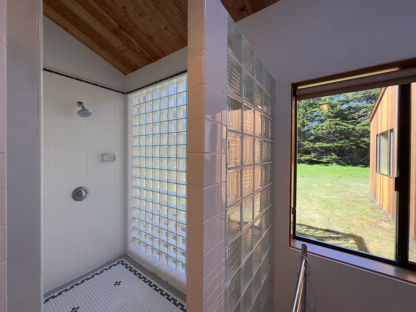 Walk-in shower with white tile, glass block partition wall, wood ceiling, and a nearby window showing an outdoor yard.