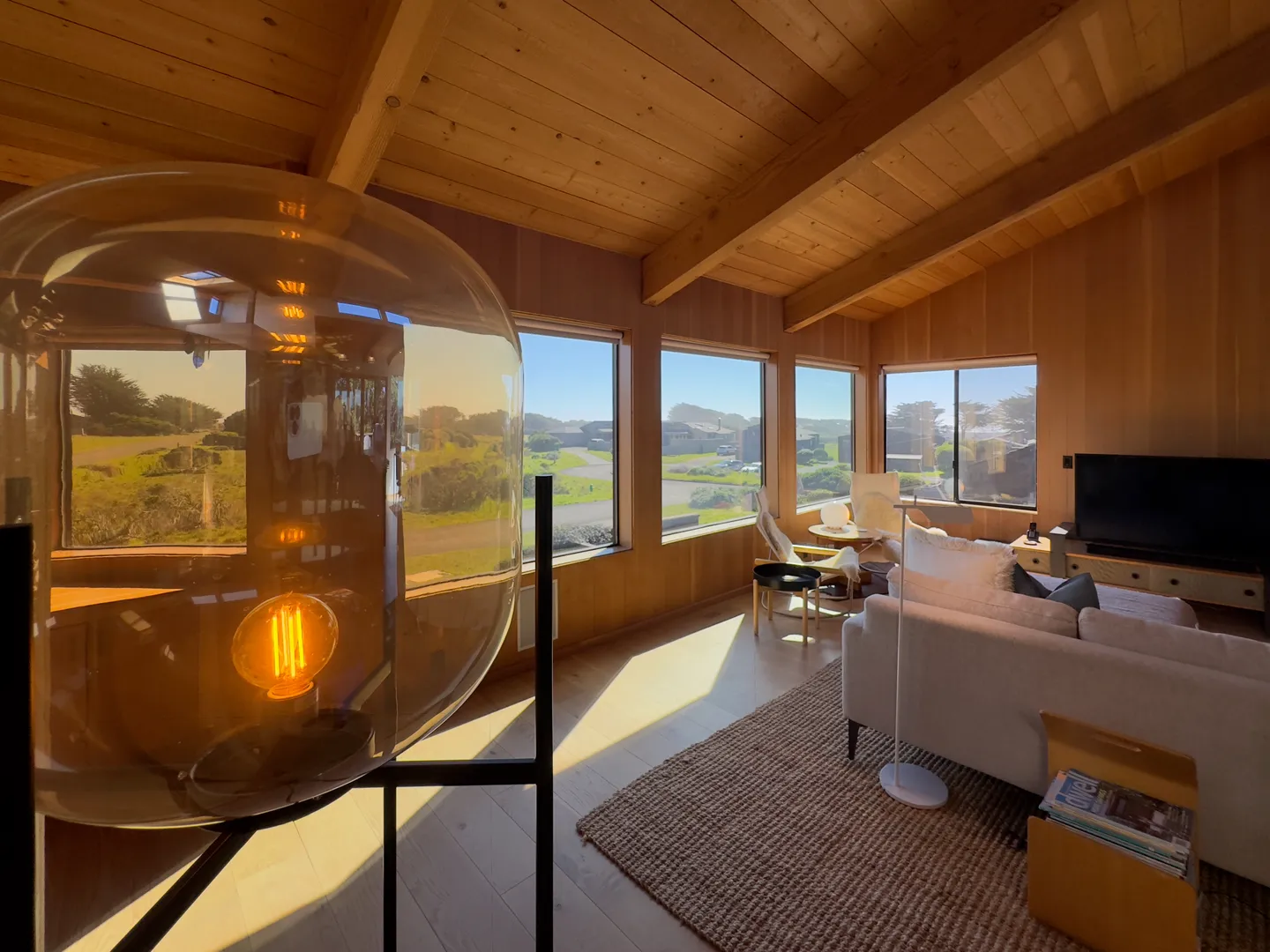 Sunlit living room with wood-paneled walls and ceiling, large windows showing an outdoor landscape, and a close-up amber glass lamp in the foreground.