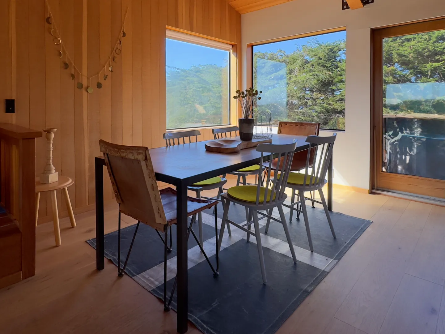 Dining area with a wooden table, chairs with green cushions, wood-paneled walls, and large windows showing green trees.