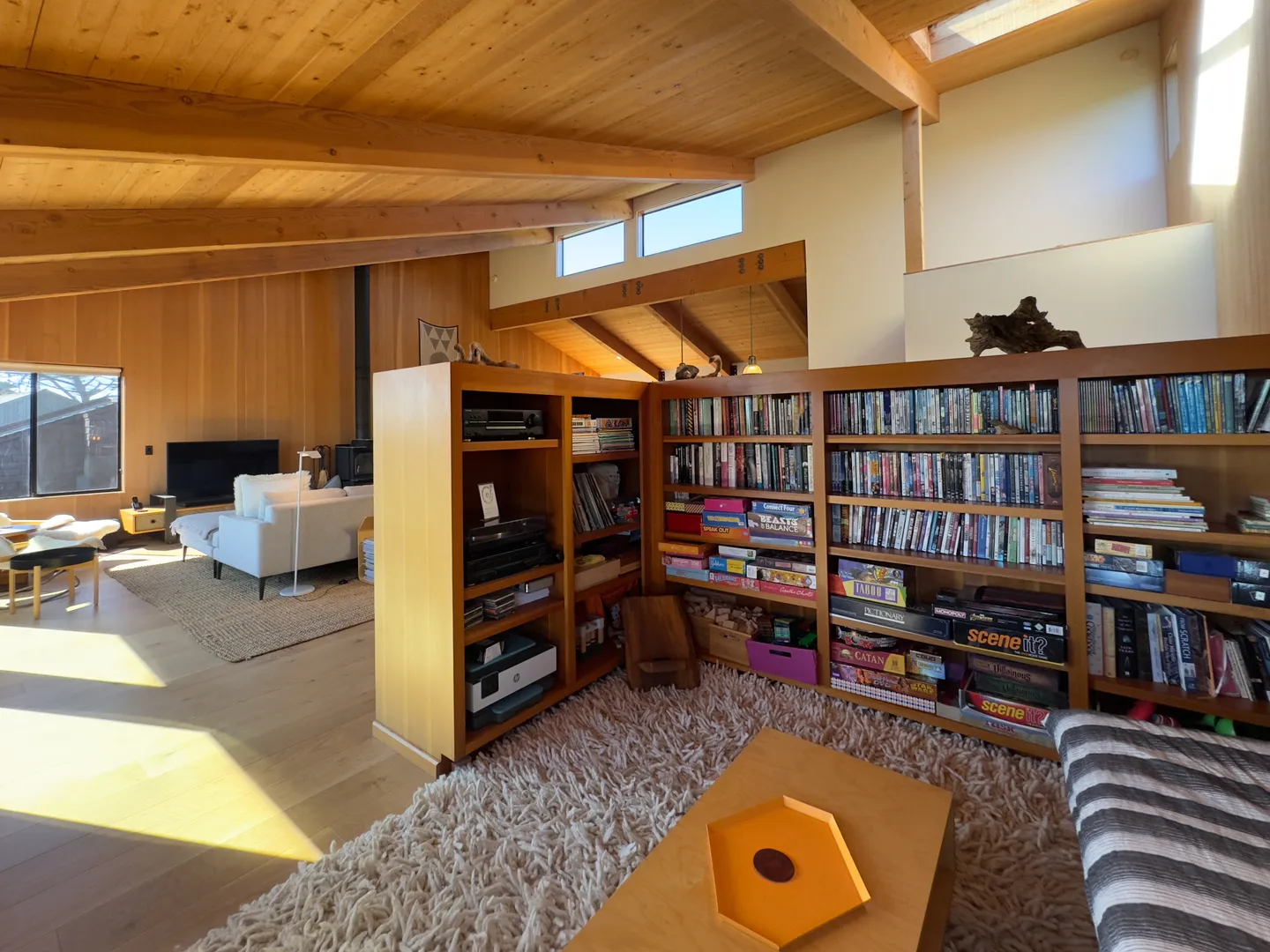 Open-concept living room and media library nook with wooden ceilings, built-in wooden bookshelves filled with games and books, and a shag rug.