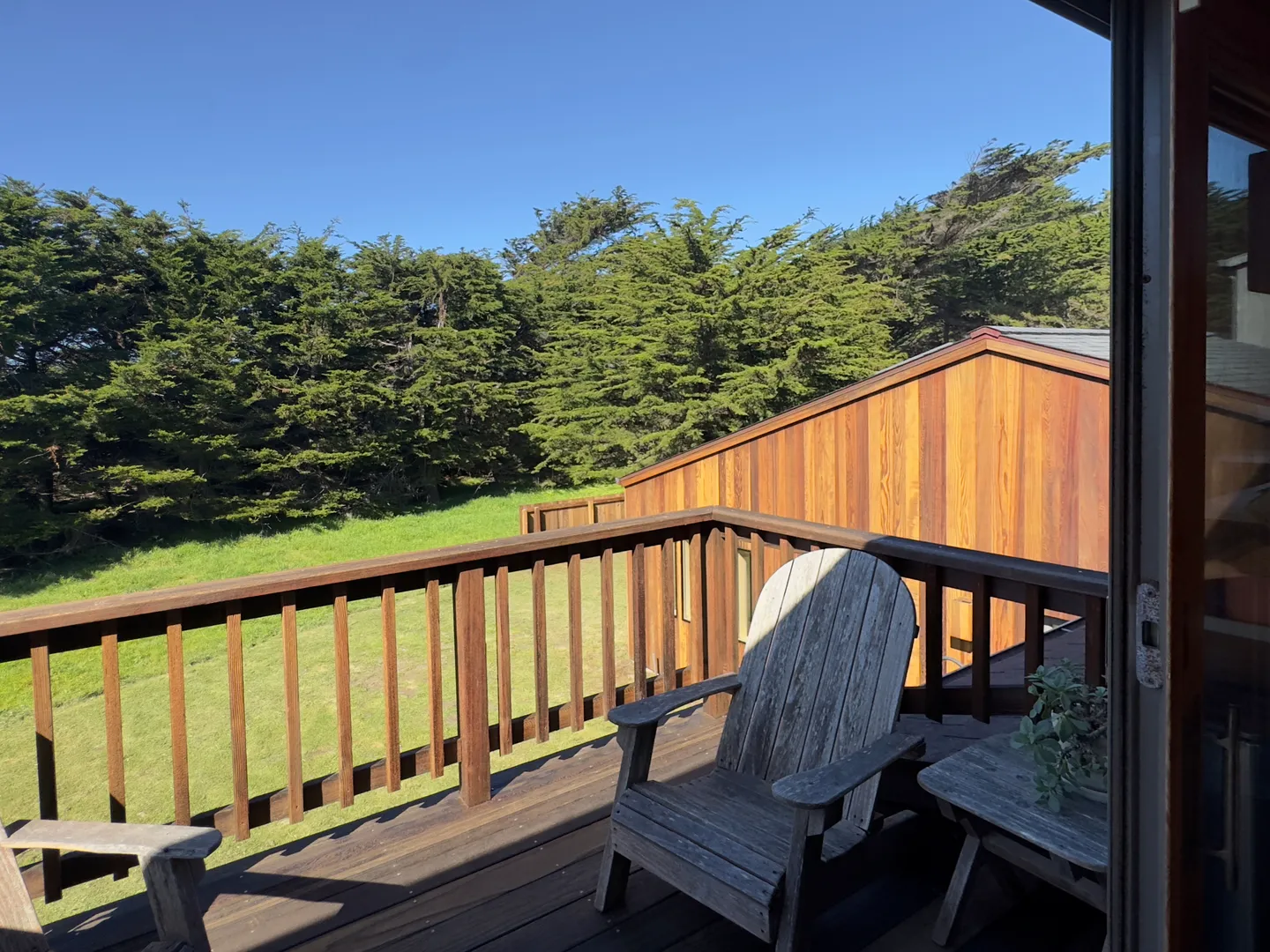 Outdoor wooden deck with railing and wooden armchairs overlooking a grassy lawn and pine trees under a clear blue sky.