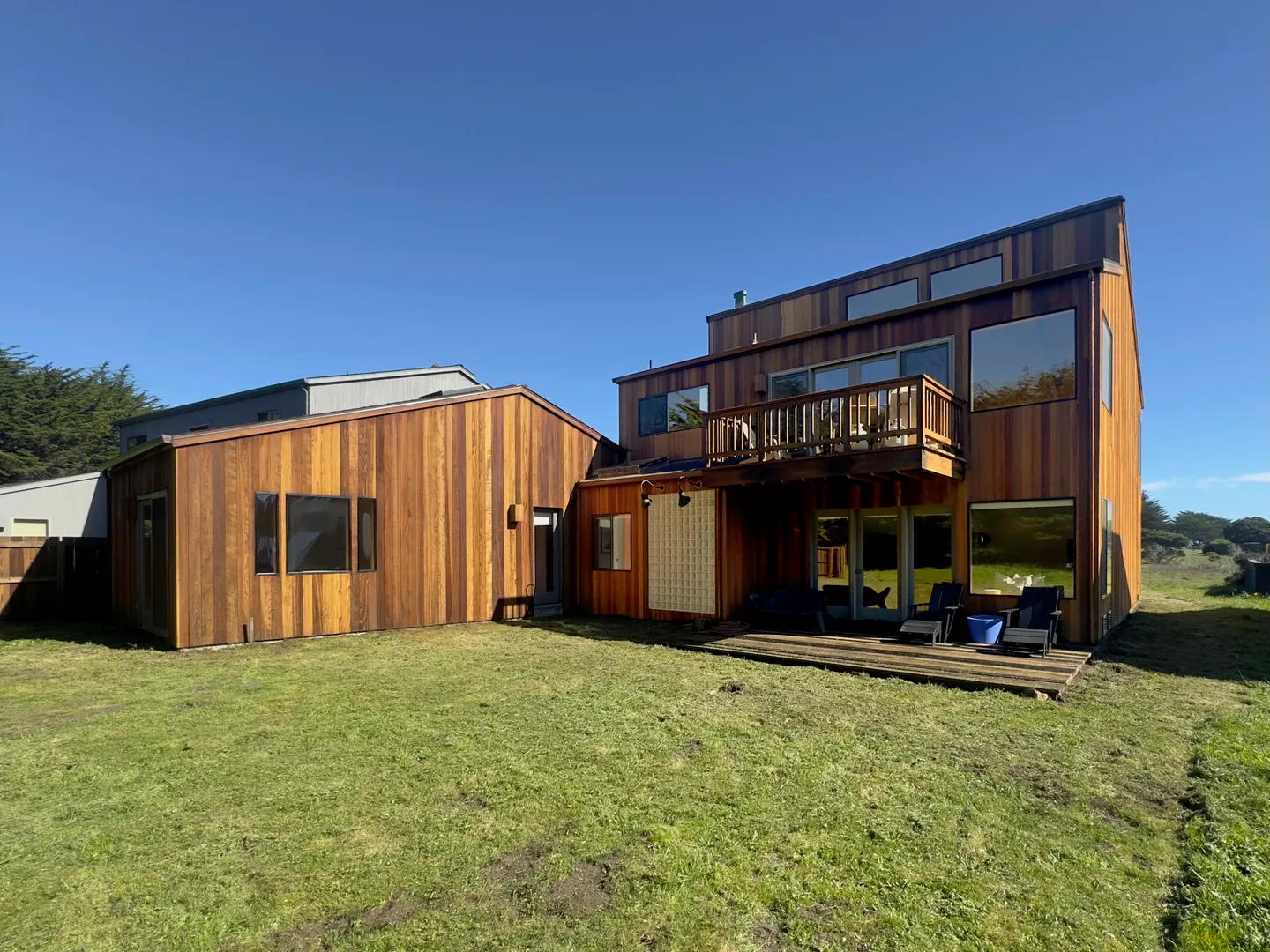 Exterior rear view of a two-story home with natural wood siding, an upper-level balcony, a wooden deck, and an expansive grassy lawn under a clear blue sky.