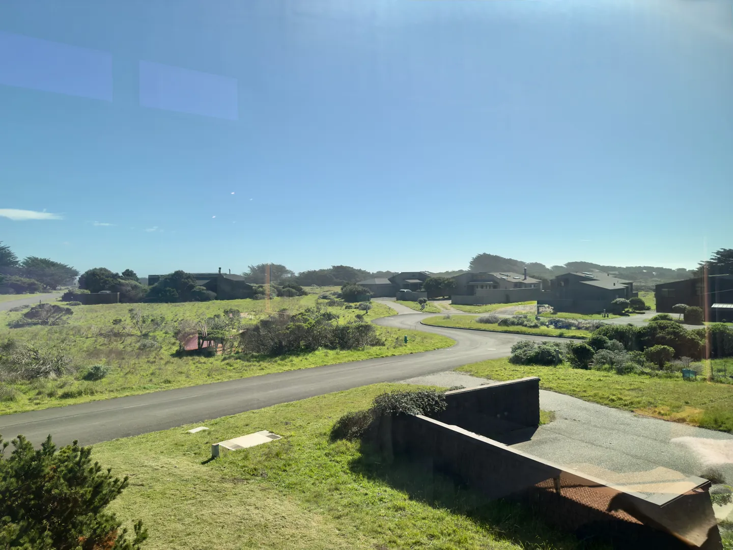 An elevated view looking through a window at a neighborhood road winding past coastal-style houses and grassy landscape under a clear blue sky.