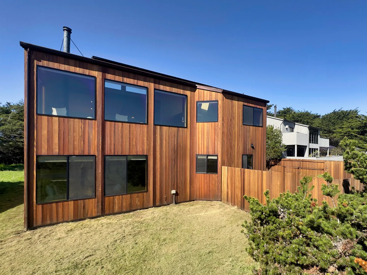 Exterior of a two-story wood-sided home with large picture windows overlooking a grassy yard under a clear blue sky.