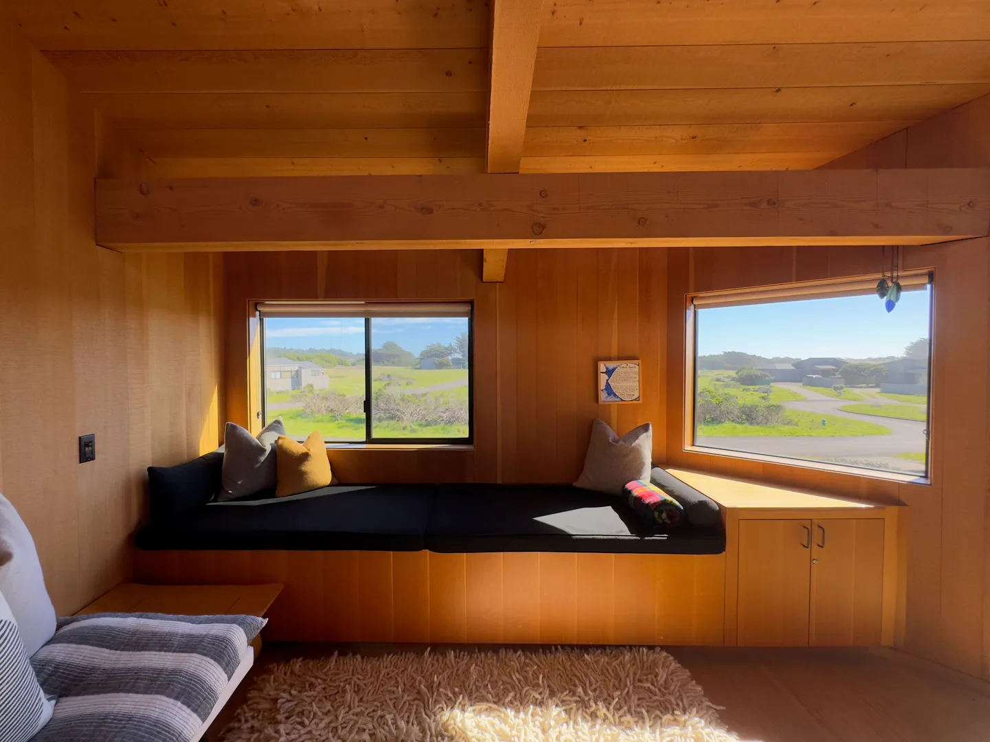 Wood-paneled room featuring a built-in daybed with cushions beneath two picture windows overlooking a coastal landscape.